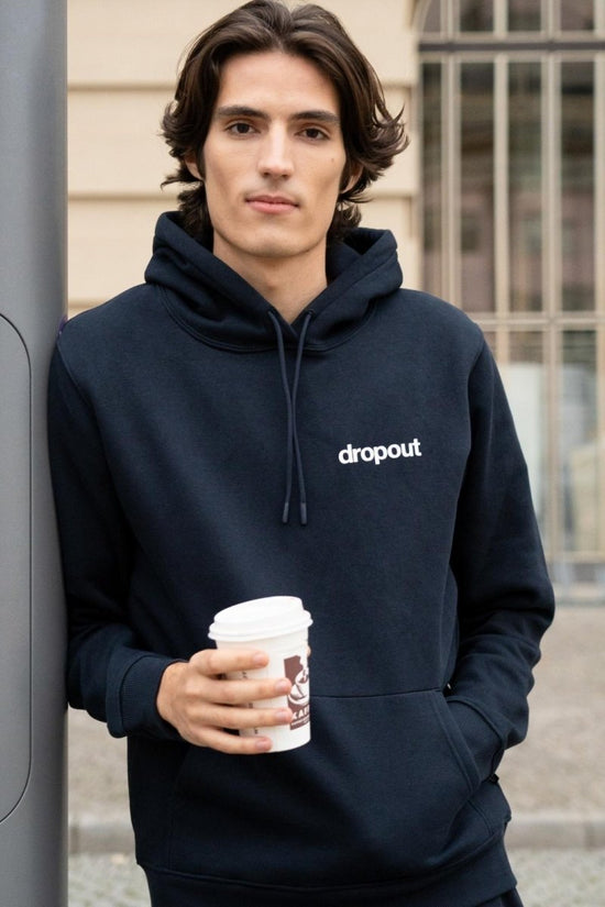 Man wearing a navy hoodie with 'dropout' logo, holding a coffee cup outdoors.