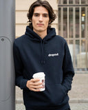 Man wearing a navy hoodie with 'dropout' logo, holding a coffee cup outdoors.