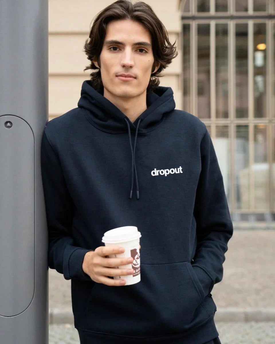 Man wearing a navy hoodie with 'dropout' logo, holding a coffee cup outdoors.