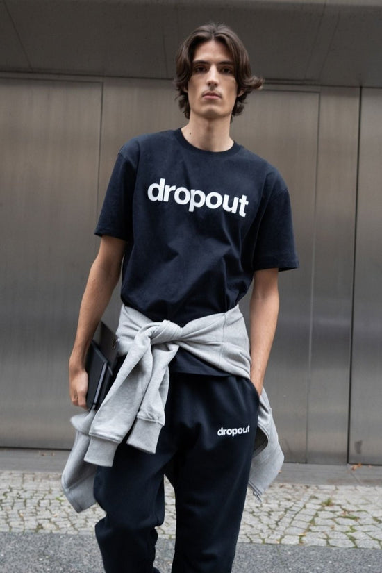 Person wearing a navy 'dropout' t-shirt with a gray sweater over one shoulder against a metallic wall.