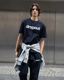 Person wearing a navy 'dropout' t-shirt with a gray sweater over one shoulder against a metallic wall.