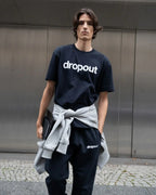 Person wearing a navy 'dropout' t-shirt with a gray sweater over one shoulder against a metallic wall.