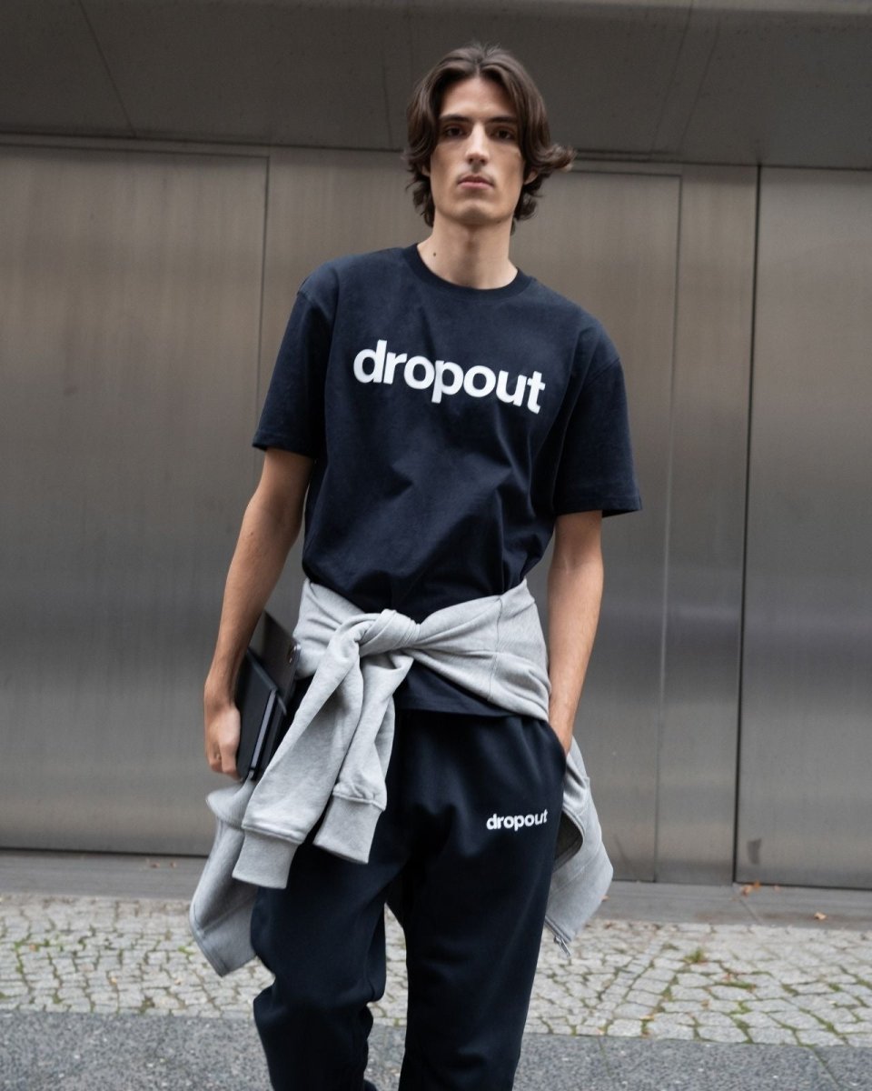 Person wearing a navy 'dropout' t-shirt with a gray sweater over one shoulder against a metallic wall.