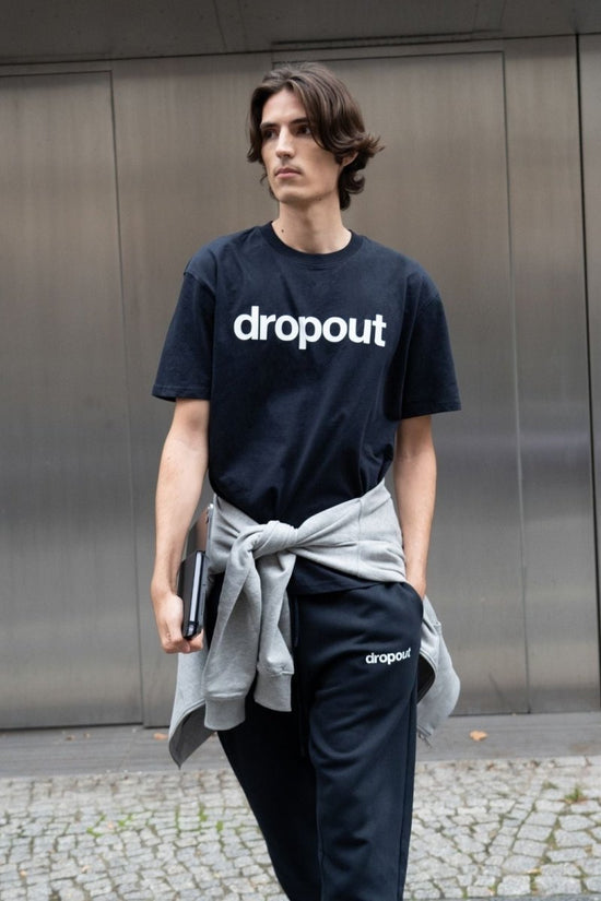 Person wearing a navy 'dropout' t-shirt and pants standing against a metallic wall.