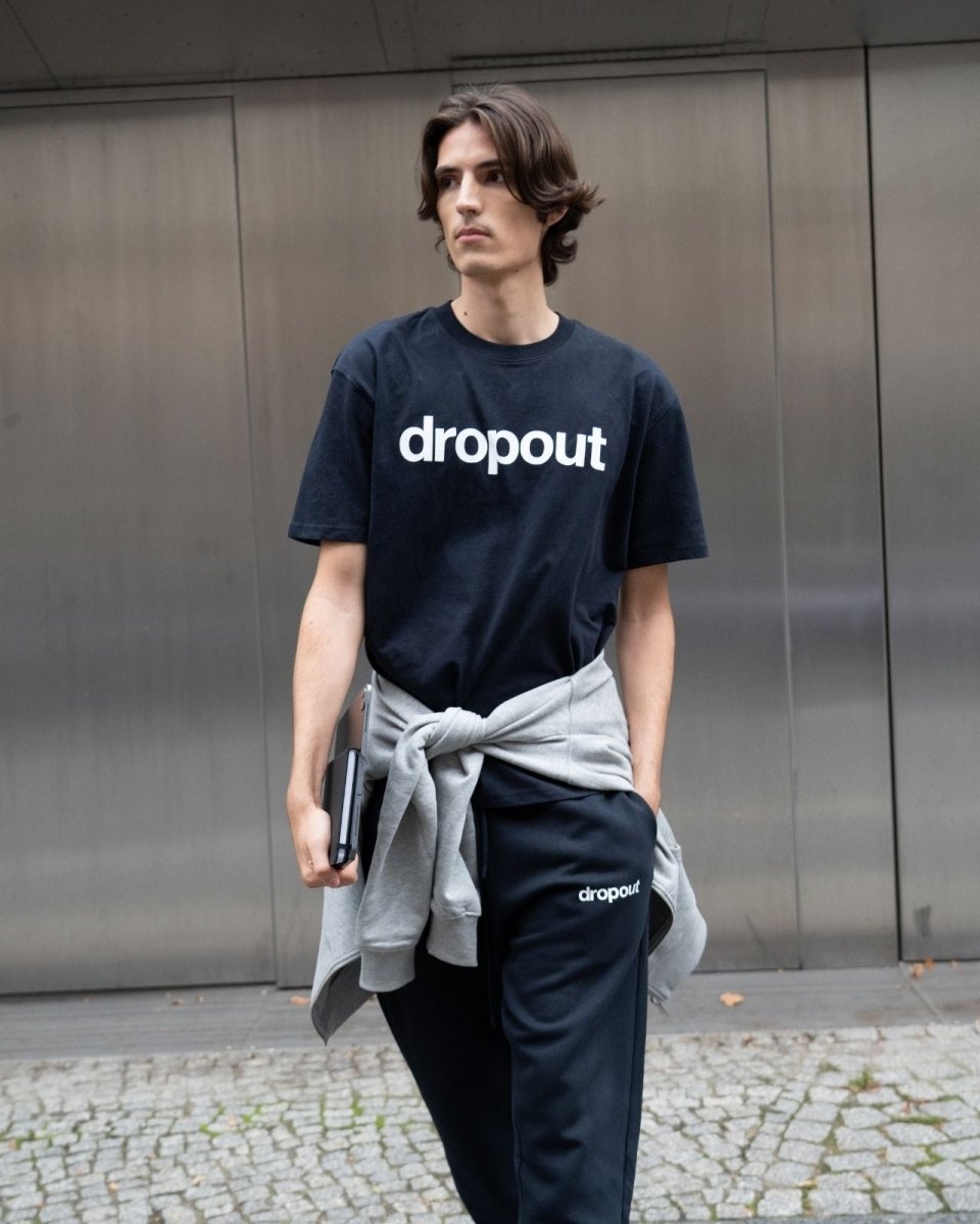 Person wearing a navy 'dropout' t-shirt and pants standing against a metallic wall.