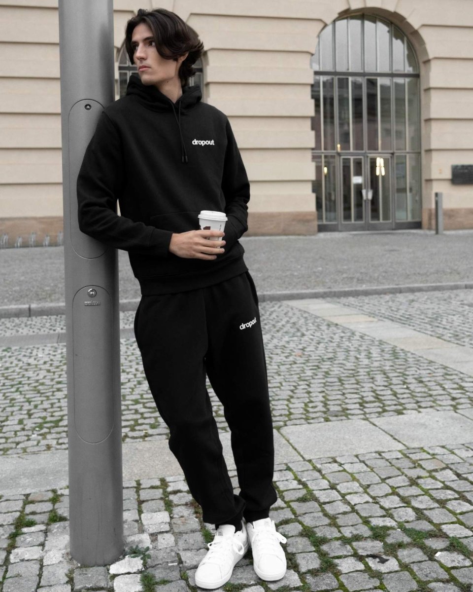 Person wearing a black hoodie with 'dropout' logo and back sweatpants, holding a coffee cup, standing on a street.