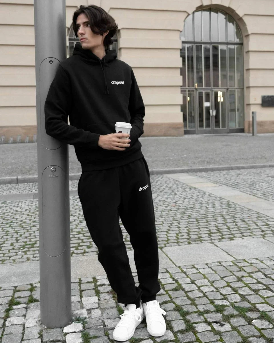 Person wearing a black hoodie with 'dropout' logo and back sweatpants, holding a coffee cup, standing on a street.