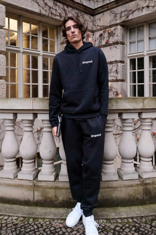 Person wearing a navy hoodie and navy sweatpants with 'dropout' branding in front of a stone building.