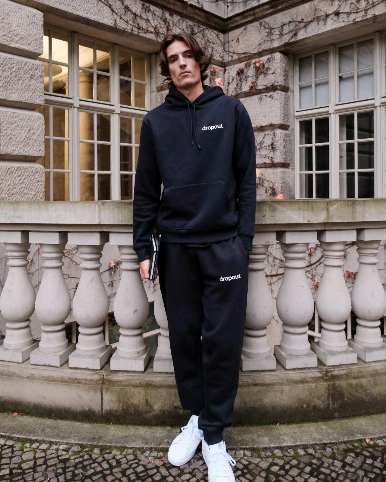 Person wearing a navy hoodie and navy sweatpants with 'dropout' branding in front of a stone building.