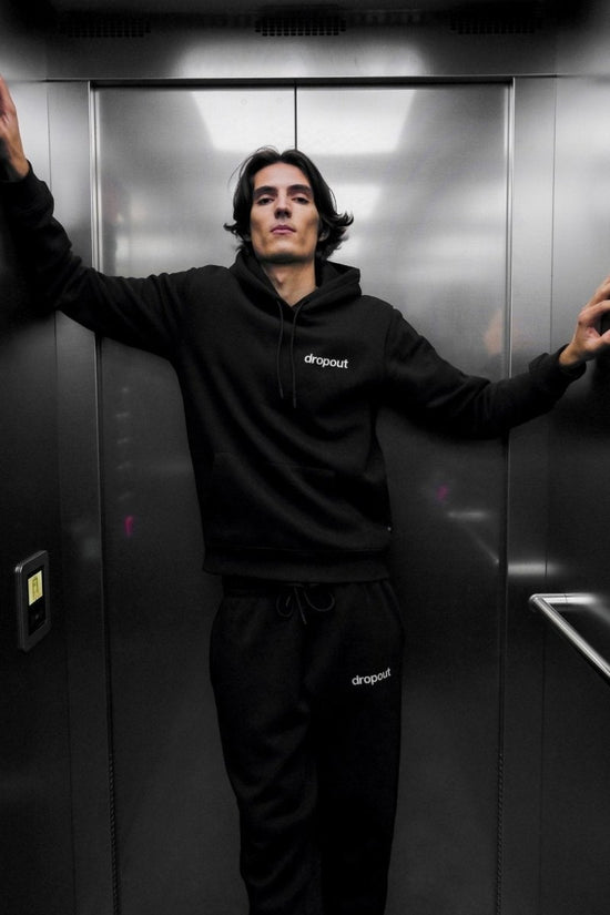 Person wearing a black hoodie and black sweatpants with 'dropout' logo in an elevator