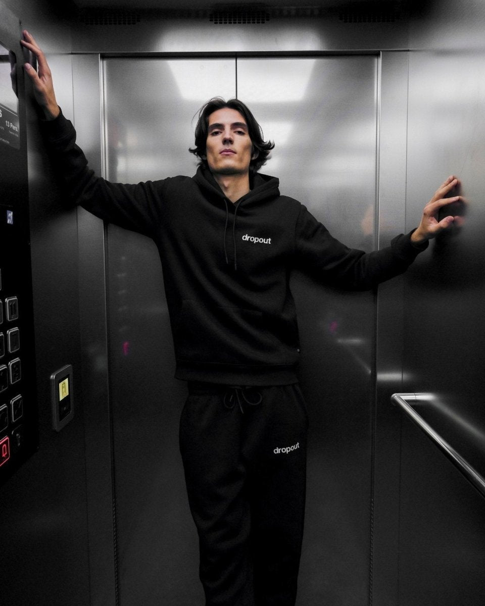 Person wearing a black hoodie and black sweatpants with 'dropout' logo in an elevator