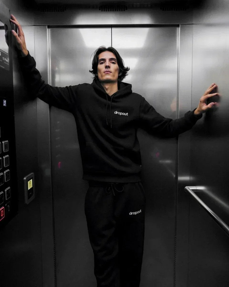 Person wearing a black hoodie and black sweatpants with 'dropout' logo in an elevator