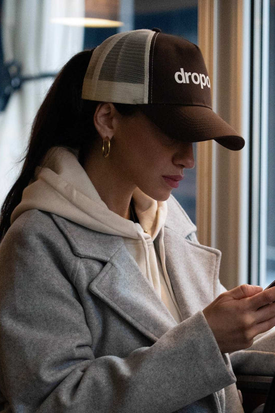 Person wearing a Brown/Beige cap with 'dropout' logo, using a phone in an indoor setting.