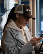 Person wearing a Brown/Beige cap with 'dropout' logo, using a phone in an indoor setting.