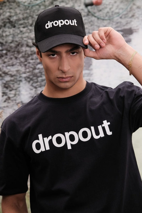 Person wearing a black 'dropout' cap and t-shirt against a textured wall.