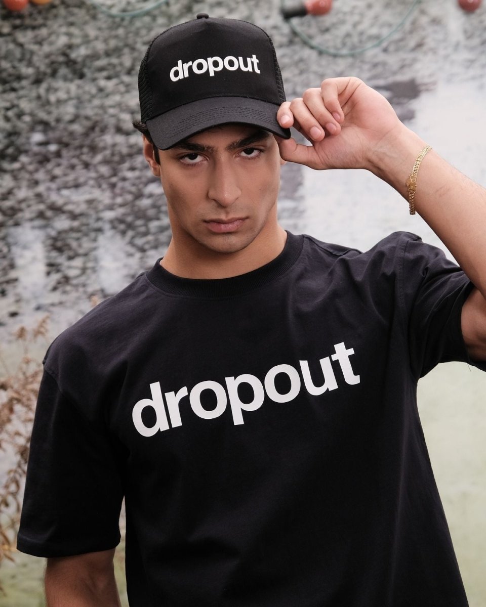 Person wearing a black 'dropout' cap and t-shirt against a textured wall.