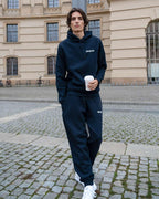Person wearing a navy hoodie and navy sweatpants with 'dropout' logo, holding a coffee cup, standing on a cobblestone street.