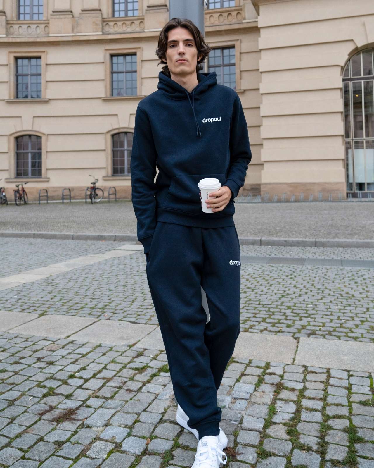 Person wearing a navy hoodie and navy sweatpants with 'dropout' logo, holding a coffee cup, standing on a cobblestone street.
