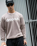 Person wearing a taupe 'dropout' sweatshirt and cap in front of a metallic fence.