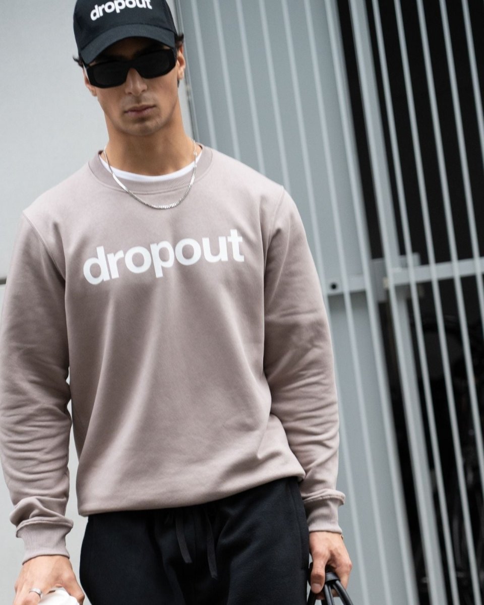 Person wearing a taupe 'dropout' sweatshirt and cap in front of a metallic fence.