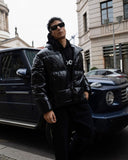 Man in a black puffer jacket standing next to a black SUV on a city street.