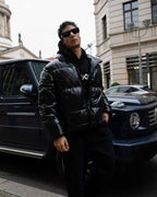 Man in a black puffer jacket standing next to a black SUV on a city street.