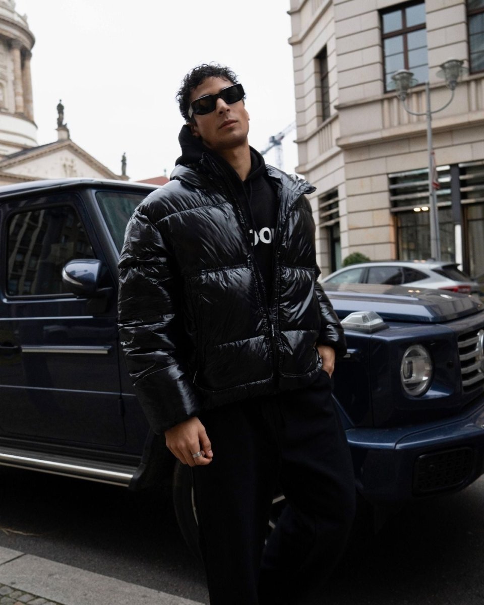 Man in a black puffer jacket standing next to a black SUV on a city street.