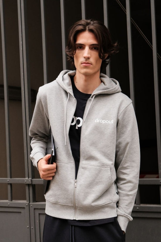 Person wearing a grey hoodie with a brand logo, standing against a metal gate background