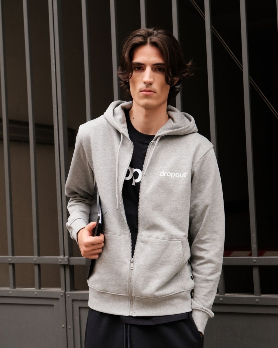 Person wearing a grey hoodie with a brand logo, standing against a metal gate background