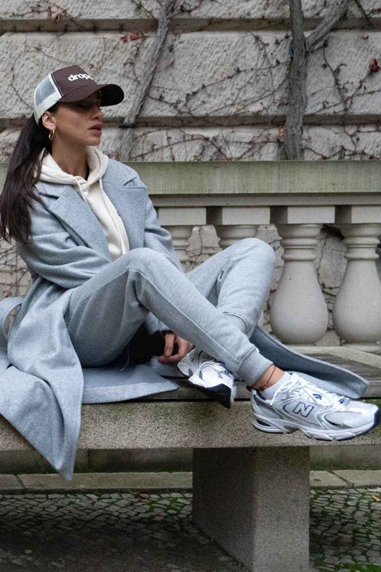 Person sitting on a bench wearing a light grey coat, grey pants, white sneakers, and a dark cap.