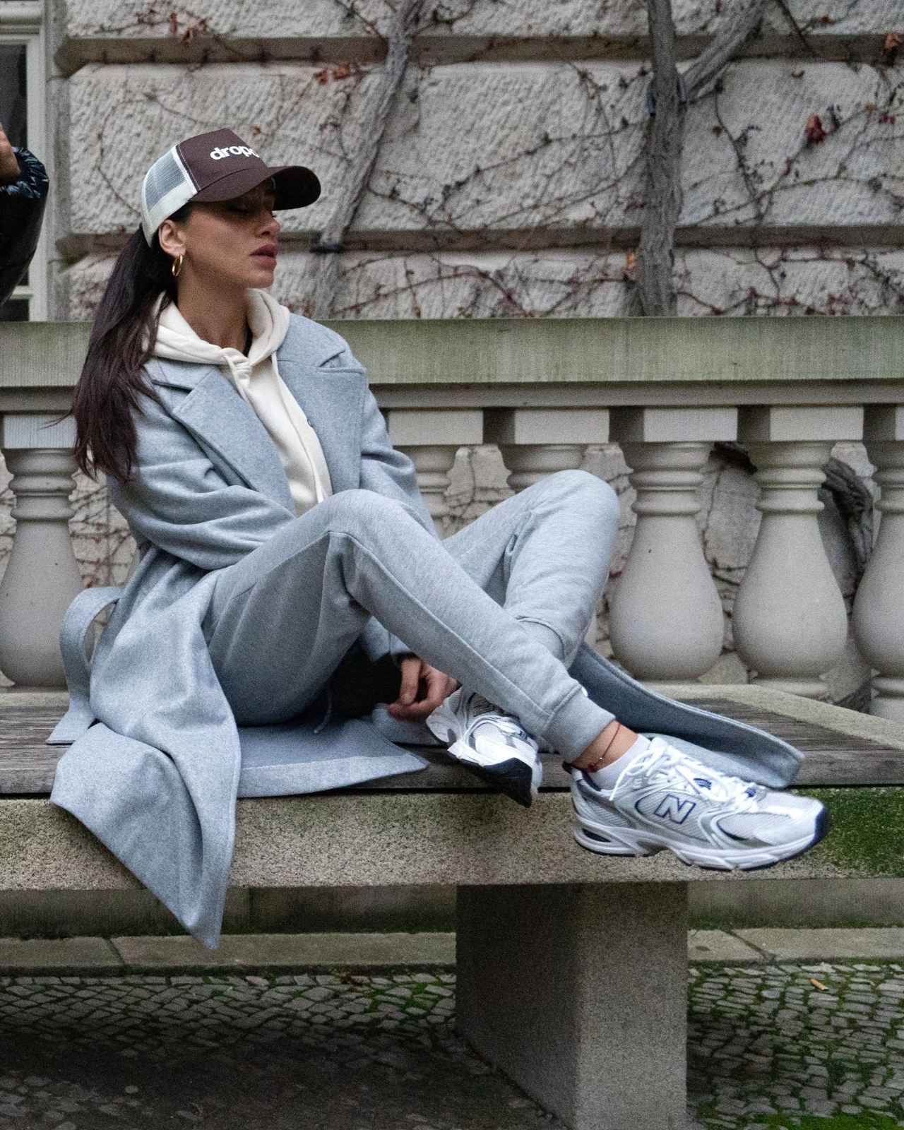 Person sitting on a bench wearing a light grey coat, grey pants, white sneakers, and a dark cap.