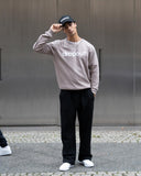 Person wearing a taupe sweatshirt with 'dropout' text, black pants, and white sneakers, standing against a metallic wall.