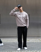 Person wearing a taupe sweatshirt with 'dropout' text, black pants, and white sneakers, standing against a metallic wall.