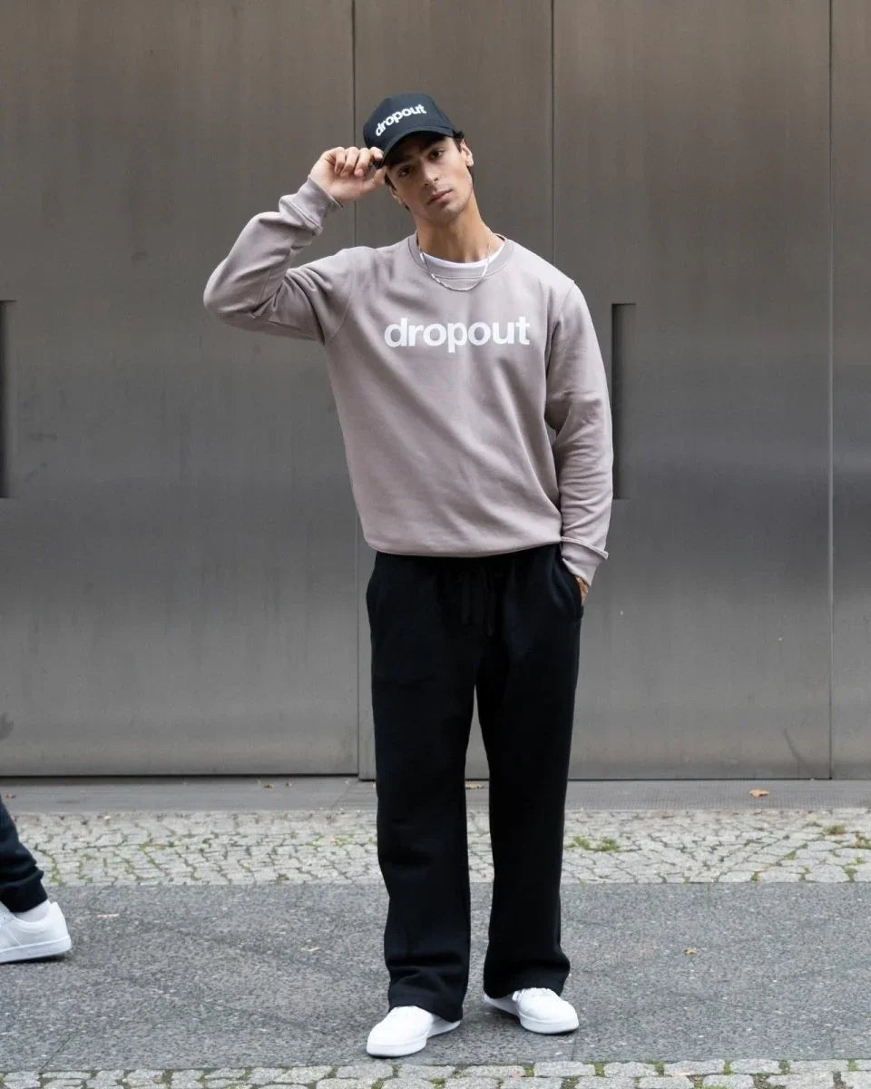 Person wearing a taupe sweatshirt with 'dropout' text, black pants, and white sneakers, standing against a metallic wall.