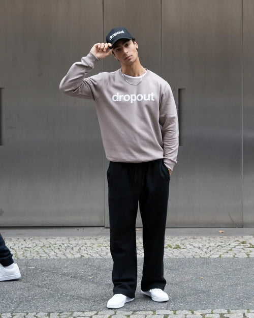 Person wearing a taupe sweatshirt with 'dropout' text, black pants, and white sneakers, standing against a metallic wall.