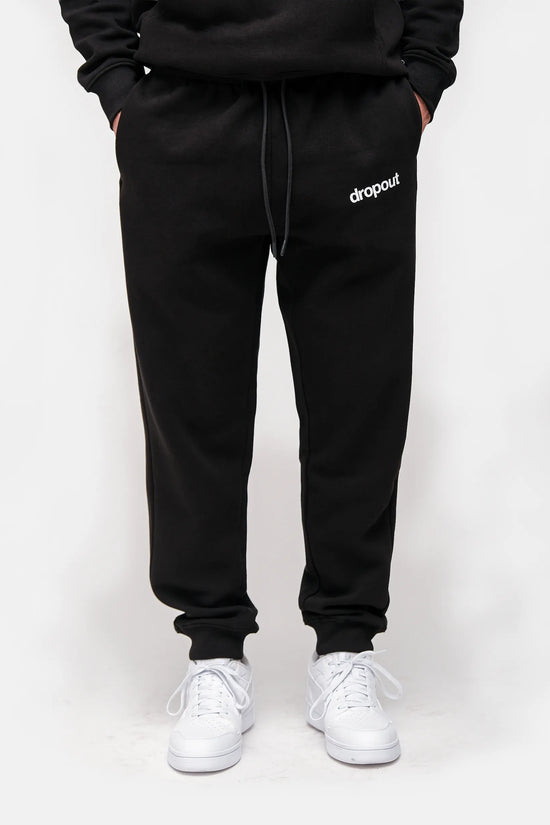 Person wearing black sweatpants with 'dropout' logo and white sneakers on a white background
