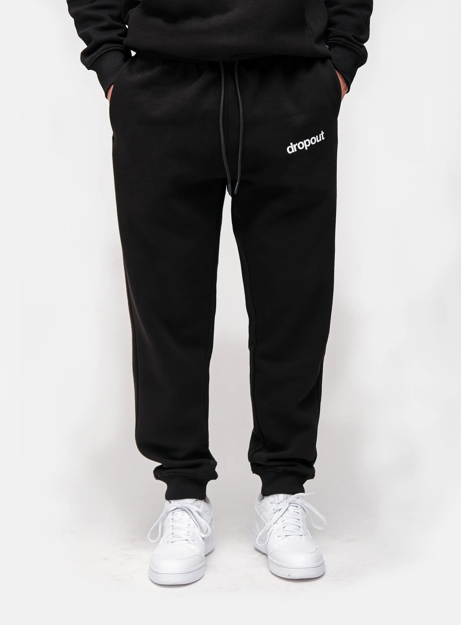 Person wearing black sweatpants with 'dropout' logo and white sneakers on a white background
