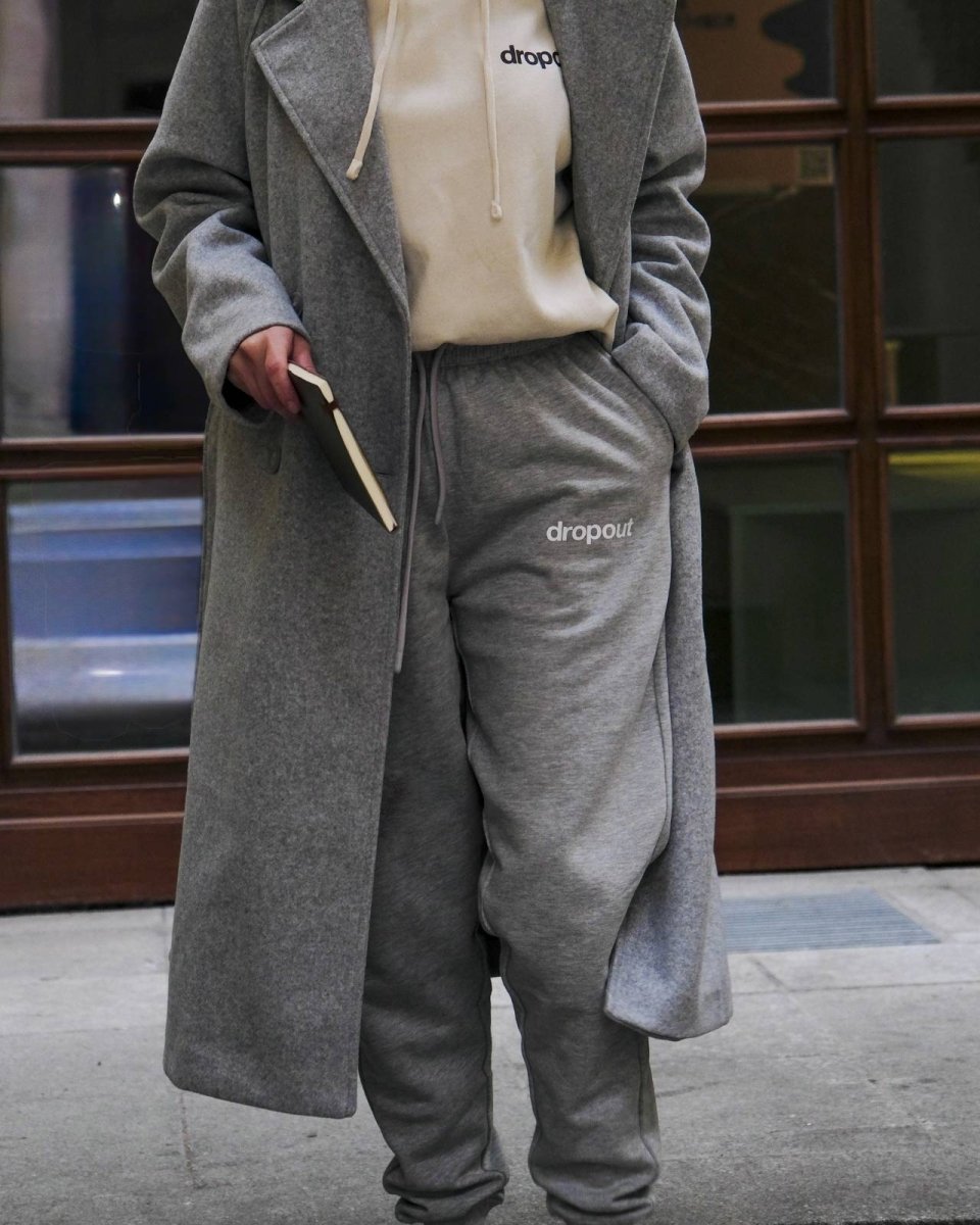 Person wearing a grey coat and matching grey sweatpants with 'dropout' branding, holding a book.