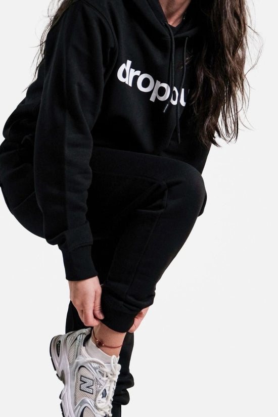Person wearing a black hoodie with 'dropout' logo and black pants on a white background