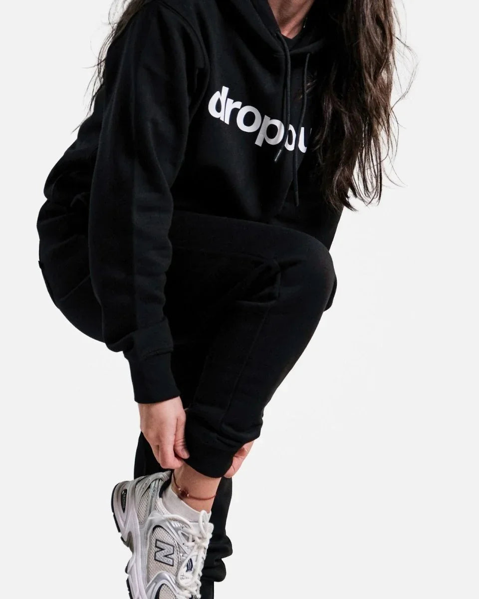 Person wearing a black hoodie with 'dropout' logo and black pants on a white background