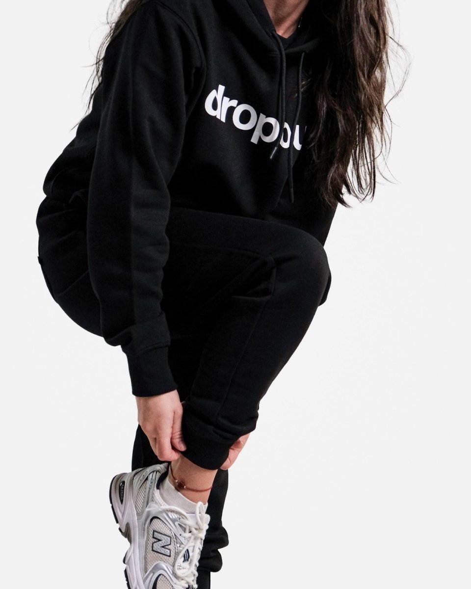 Person wearing a black hoodie with 'dropout' logo and black pants on a white background