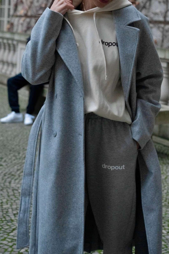 Person wearing a grey coat over a white hoodie and grey pants with 'dropout' branding.