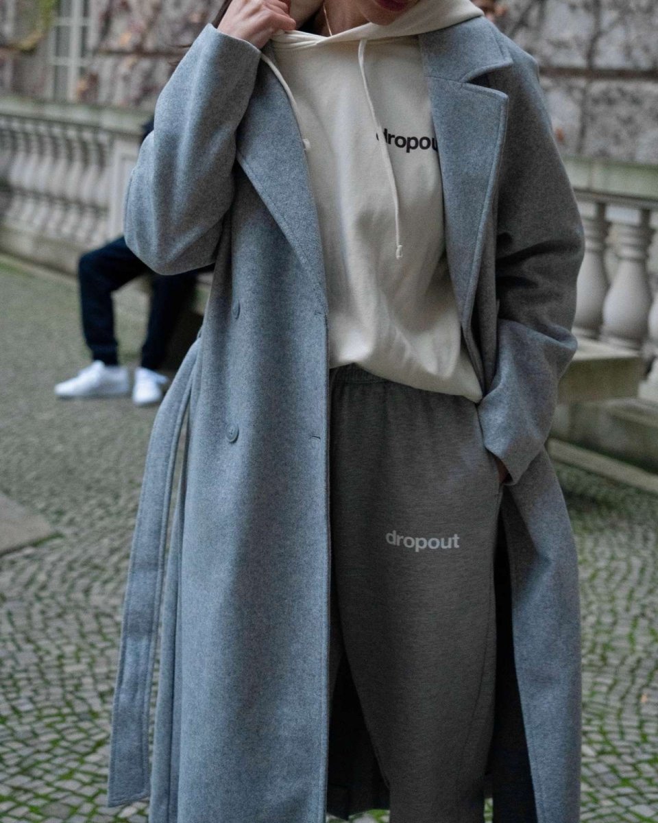 Person wearing a grey coat over a white hoodie and grey pants with 'dropout' branding.