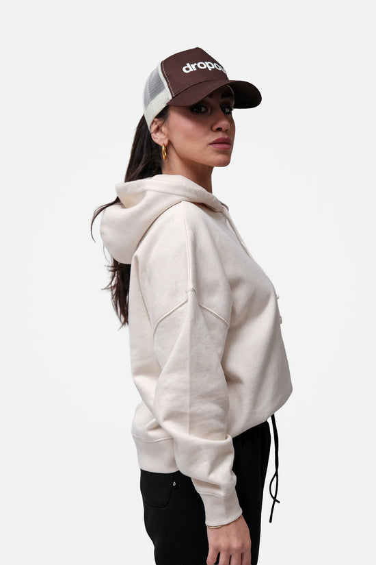 Person wearing a beige hoodie and brown cap with 'drop' logo on a white background