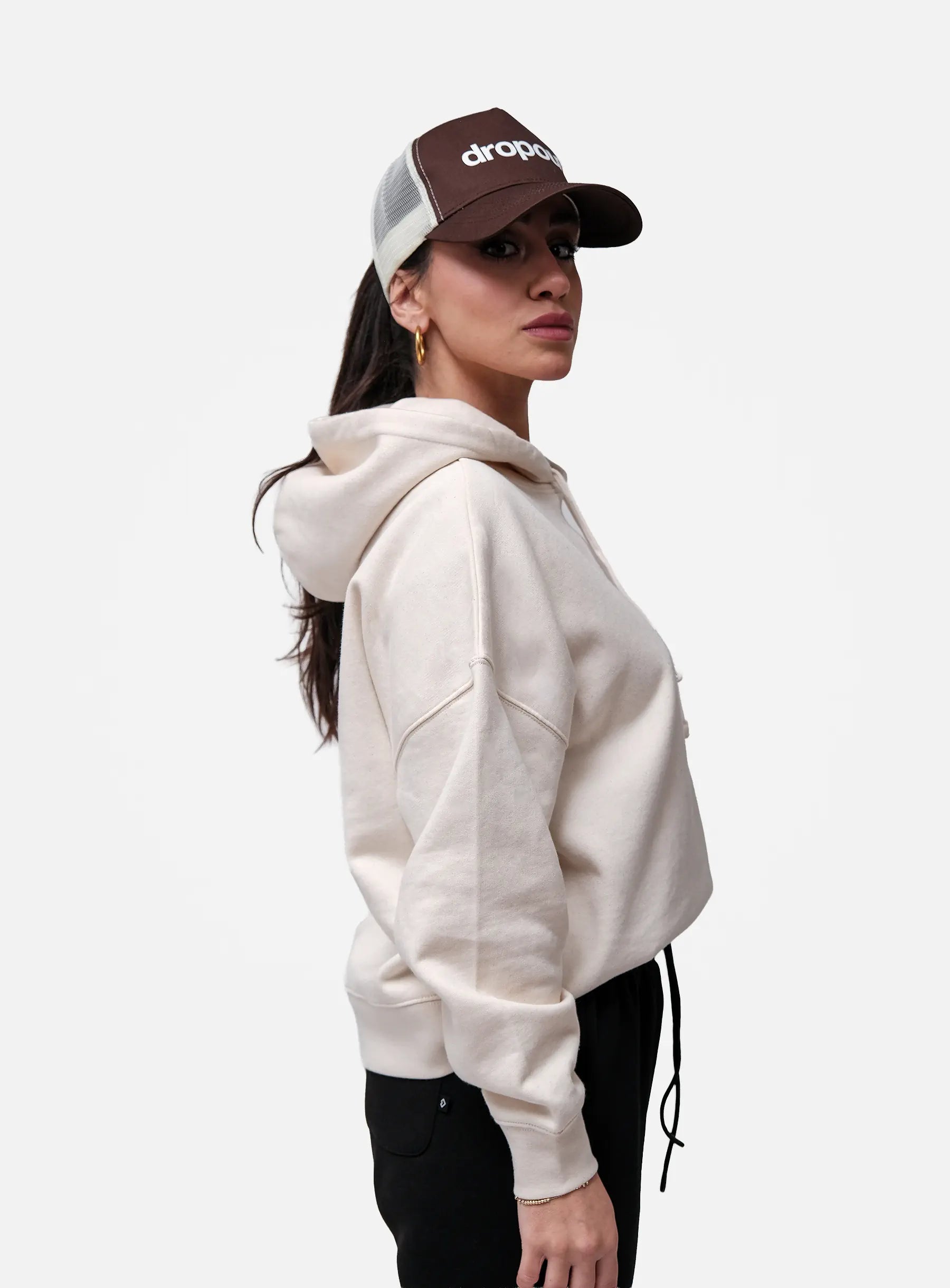 Person wearing a beige hoodie and brown cap with 'drop' logo on a white background
