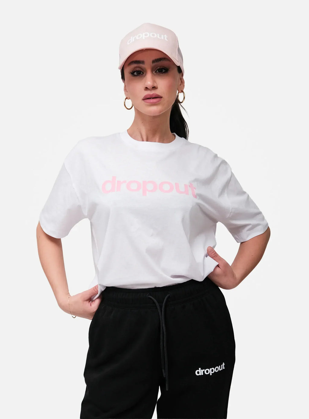 Person wearing a white 'dropout' t-shirt and black pants with a pink cap on a white background