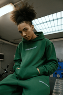 Person wearing green hoodie and sweatpants with text design, posing indoors with industrial background.