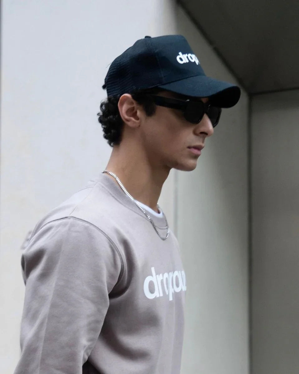 Man wearing dropout Milano beige sweatshirt and black cap, streetwear style, urban setting.