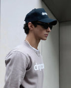 Man wearing dropout Milano beige sweatshirt and black cap, streetwear style, urban setting.