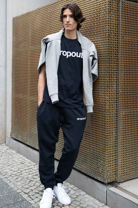 Person wearing a navy 'dropout' t-shirt and pants with a gray jacket against a textured wall.